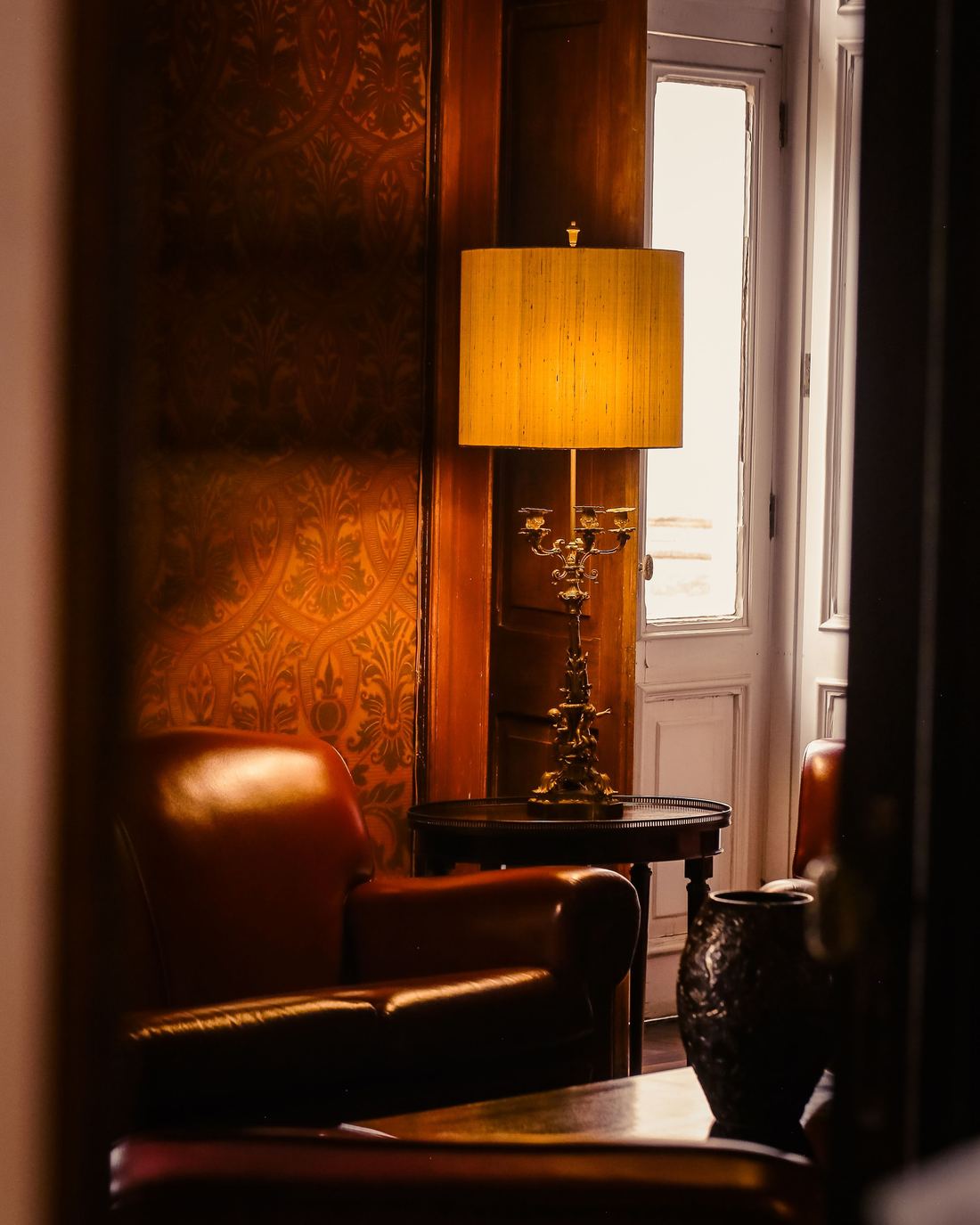 Warm sitting room corner with dark patterned wallpaper, a leather armchair and an amber-shaded lamp
