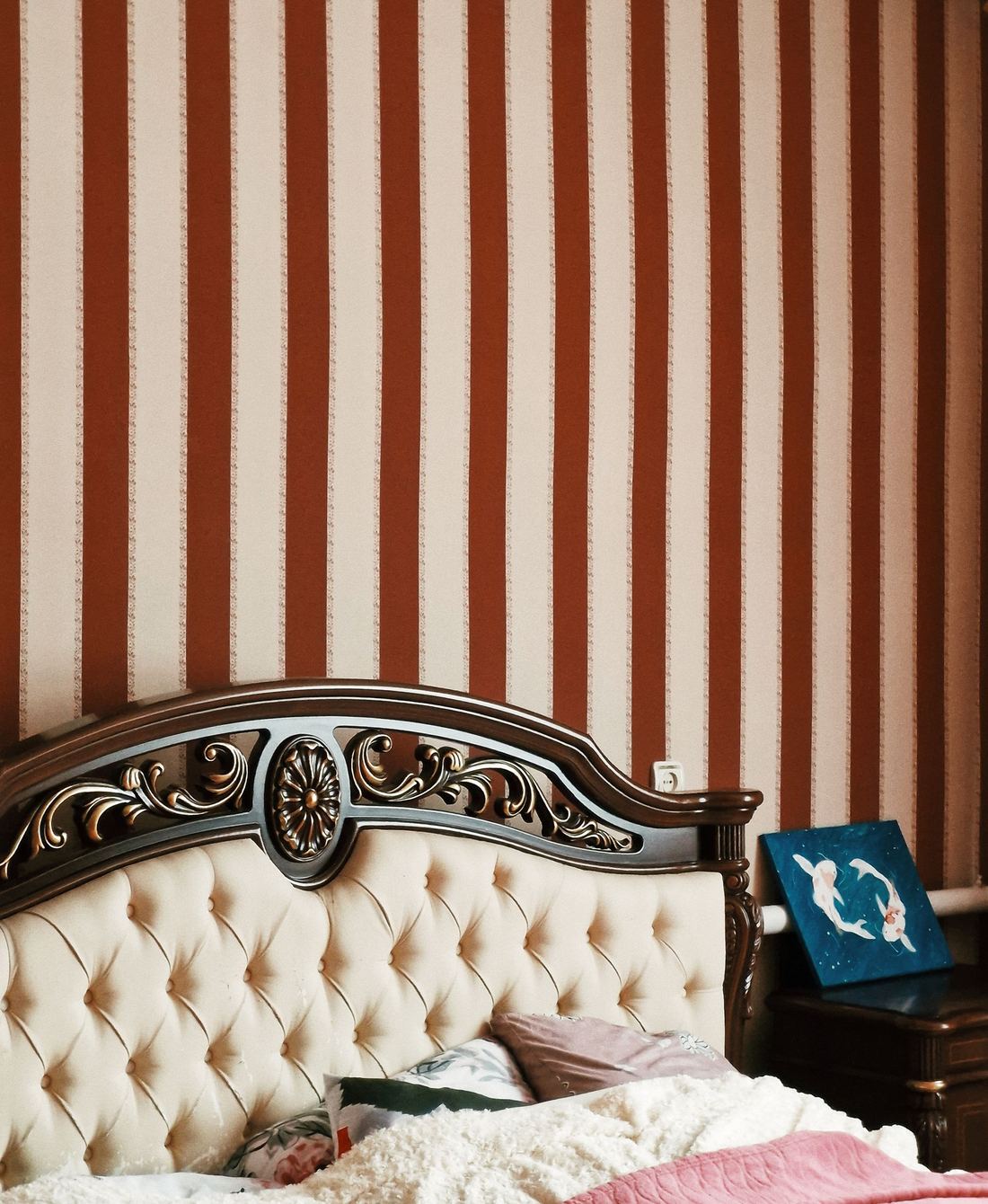 Traditional bedroom with red and ivory striped wallpaper above a tufted, carved headboard