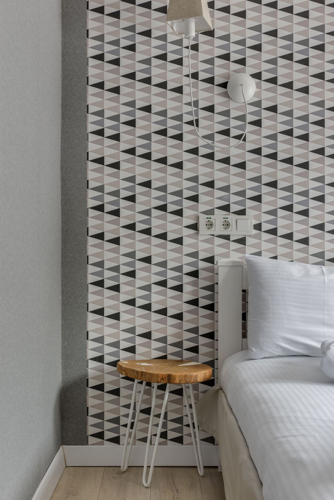 Bedroom corner with black, white and grey geometric triangle wallpaper, a white bed and a wooden stool